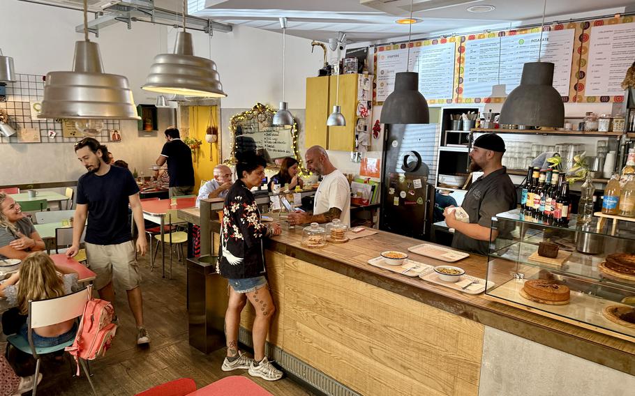 Interior view of a modern casual restaurant with an open kitchen, pendant lighting, colorful seating, menu boards on the walls and customers ordering at the counter while others dine at tables.