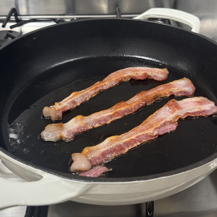 The Tramontina Bestow Enameled Cast Iron Skillet over a gas stove with bacon cooking in it.