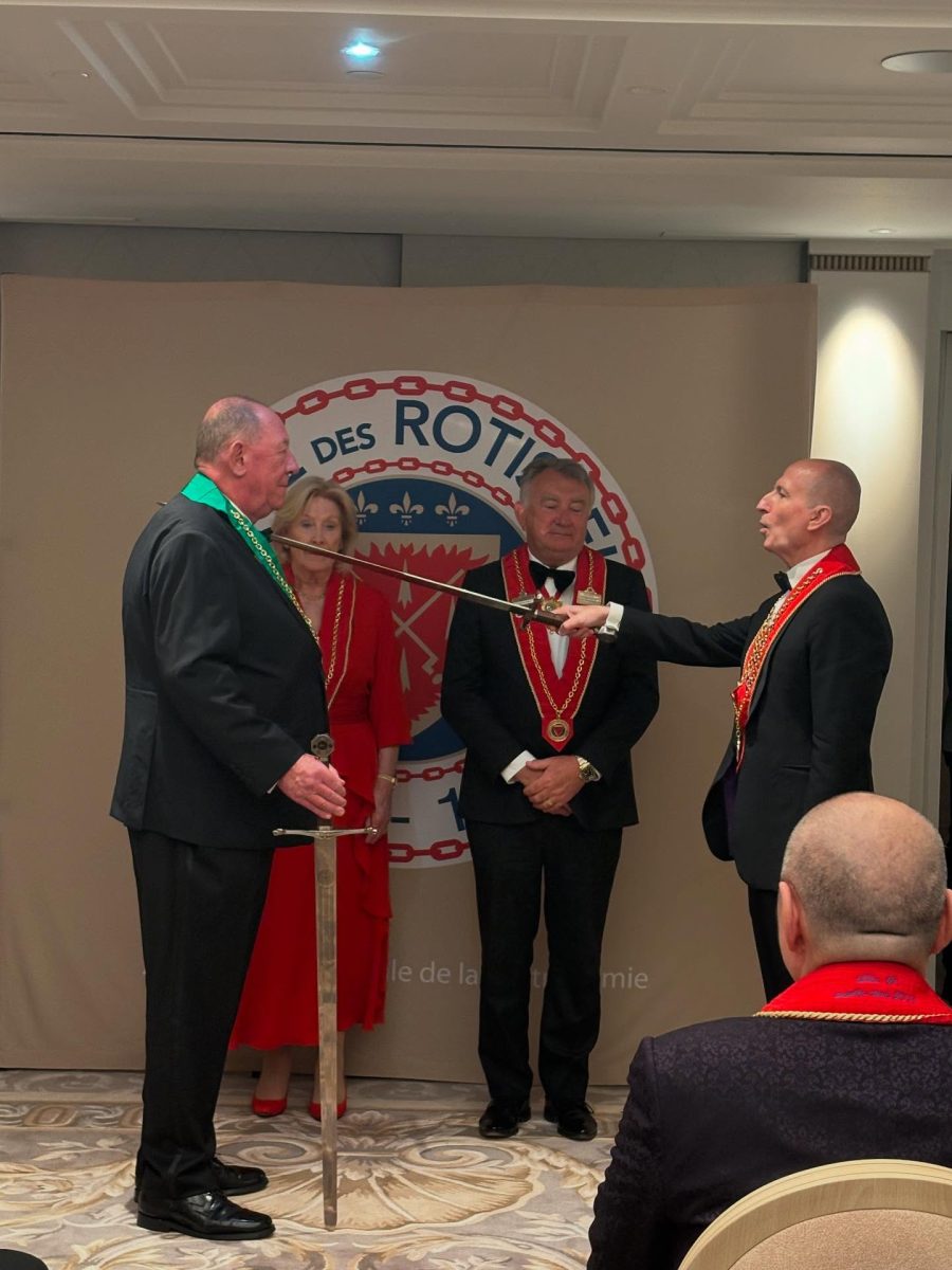 A man being dubbed with a sword by another man - both are wearing coloured ribbons with medallions, in the background two other people stand in front of the symbol of La Chaîne des Rôtisseurs. 