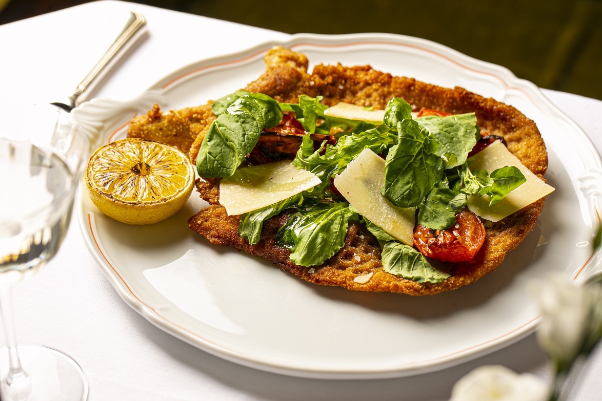 A crispy breaded dish topped with arugula, tomato, cheese, served with a grilled lemon on a white plate with cutlery.