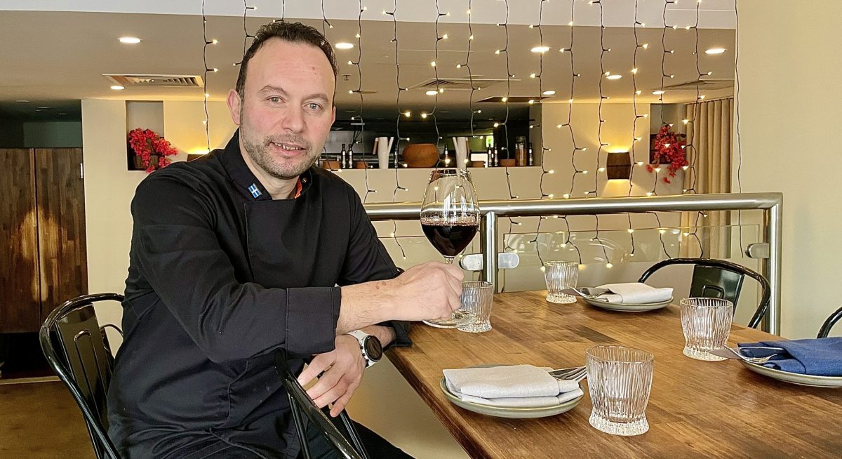 Man in black chefs uniform sits at restaurant table with a glass of red wine.