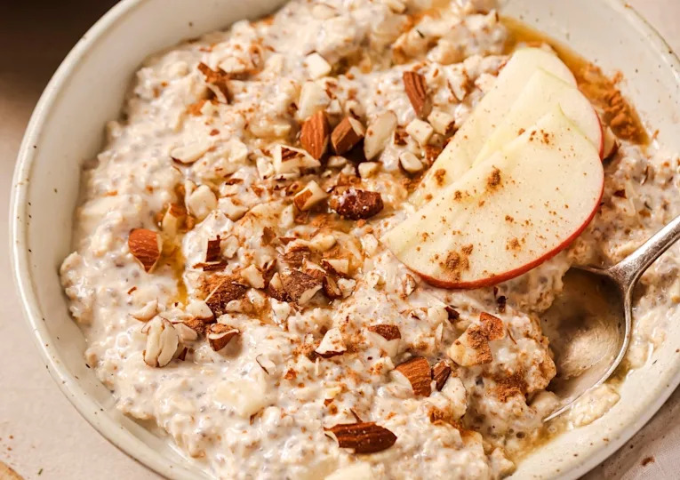 A bowl of oatmeal topped with chopped almonds, apple slices, and a drizzle of honey. A spoon is placed in the pudding.