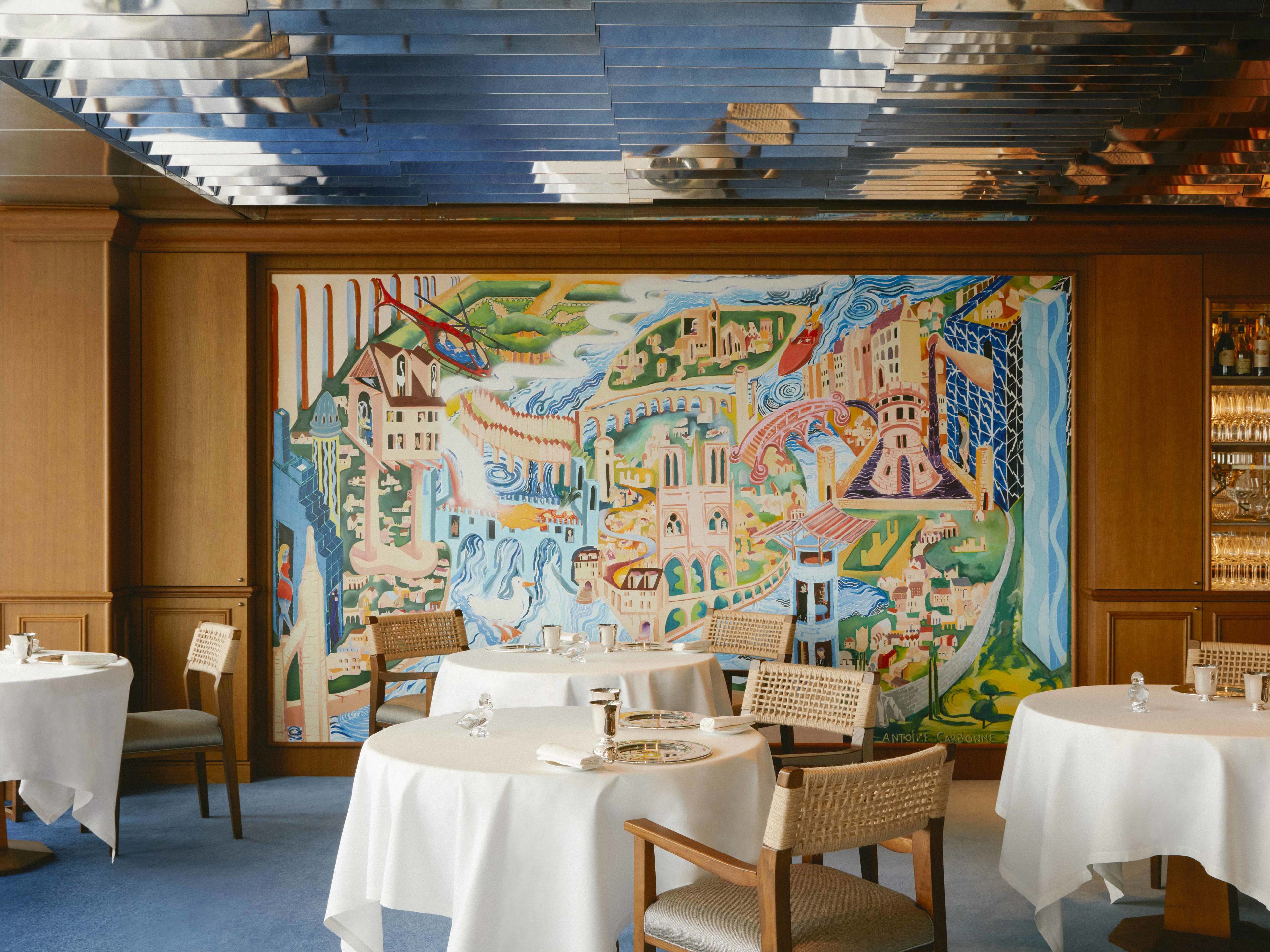 A bright dining room with a big mural in the background, and tables with white tablecloths in the foreground.