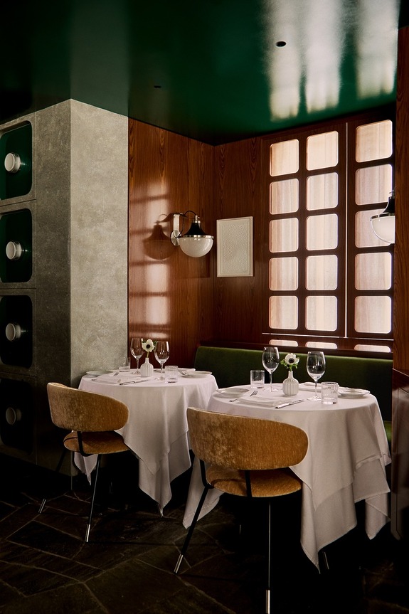 Elegant restaurant interior with wooden walls, green ceiling, and two tables set for dining with white tablecloths and modern chairs.
