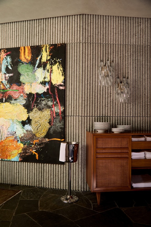 Modern interior with abstract painting, wood cabinet stocked with plates, glasses, and a sleek metal ice bucket on the floor.