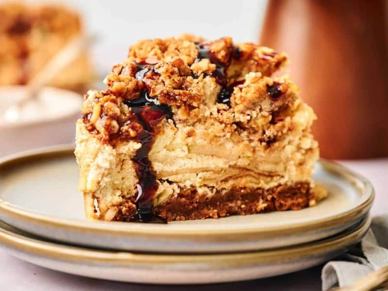 A slice of apple crumble cheesecake with a crumbly topping and a drizzle of dark sauce, served on a ceramic plate.