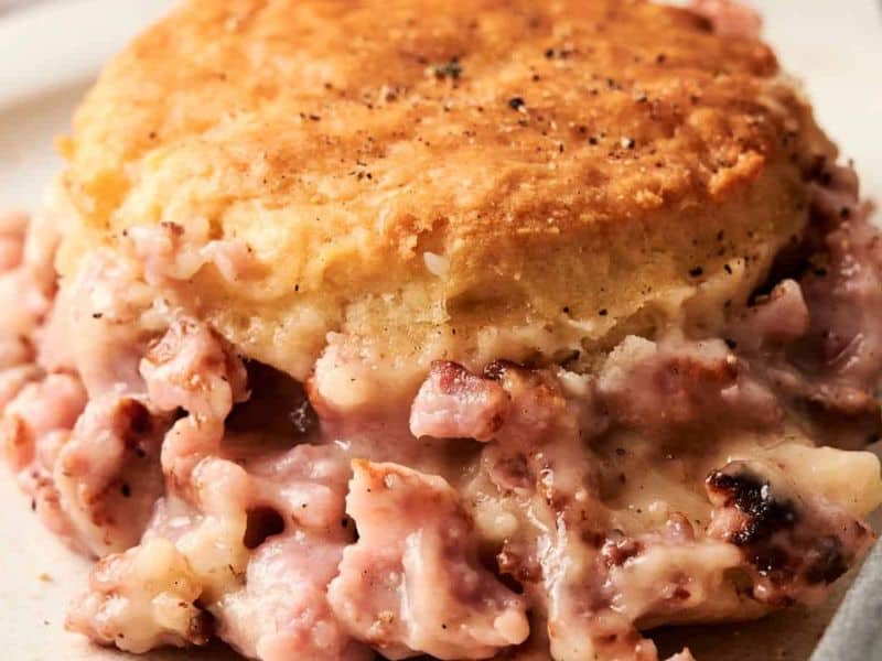 A close-up of a biscuit topped with creamy sausage gravy, featuring visible chunks of sausage and a golden-brown biscuit.