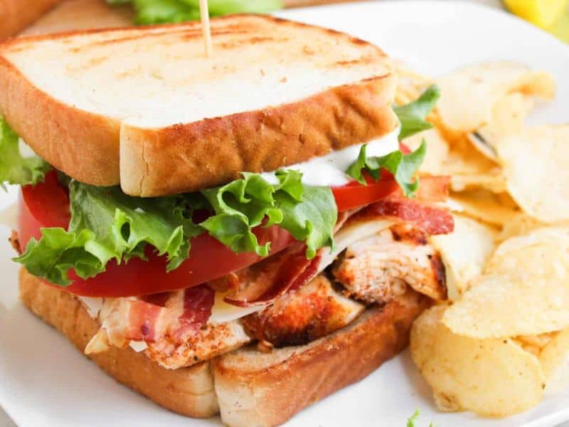 A grilled chicken sandwich with lettuce, tomato, bacon, and mayonnaise on toasted bread, served with potato chips on a white plate.