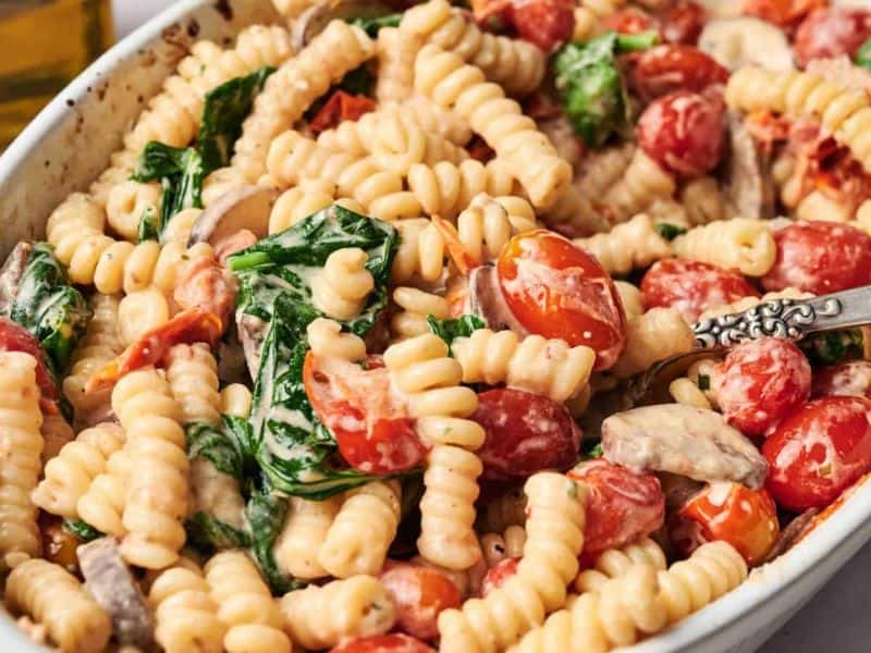 A close-up of cooked pasta mixed with cherry tomatoes, spinach, mushrooms, and a creamy sauce in a white dish with a spoon.