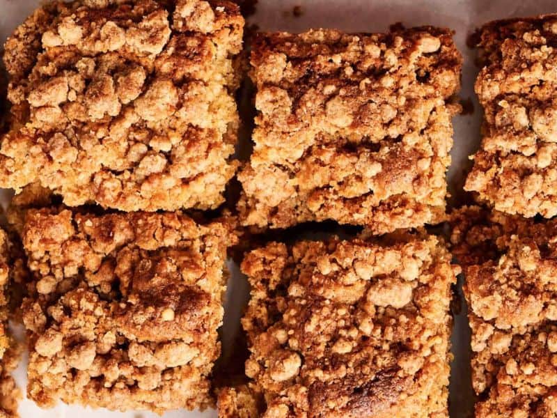 Six pieces of crumb-topped coffee cake sit close together on a white surface, showing a golden brown, crumbly texture.