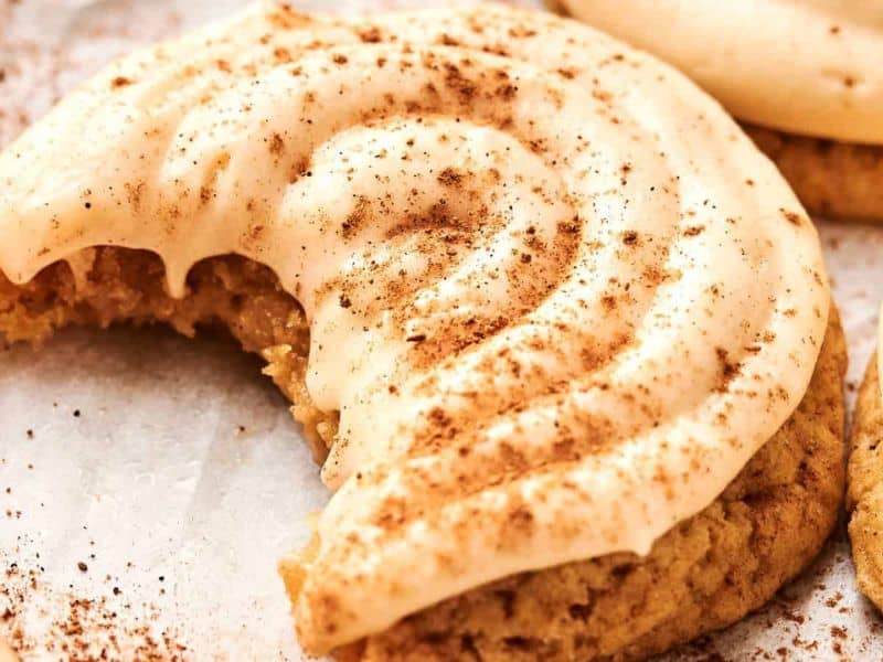 A frosted cookie with a bite taken out, topped with cream-colored icing and sprinkled with cinnamon, rests on a light-colored surface.