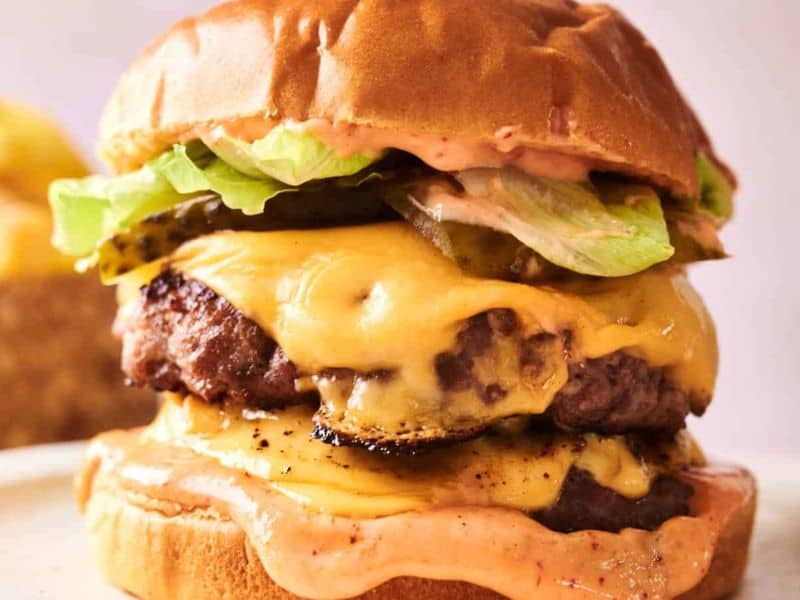 Close-up of a cheeseburger with two beef patties, melted cheese, lettuce, pickles, and sauce on a toasted bun.