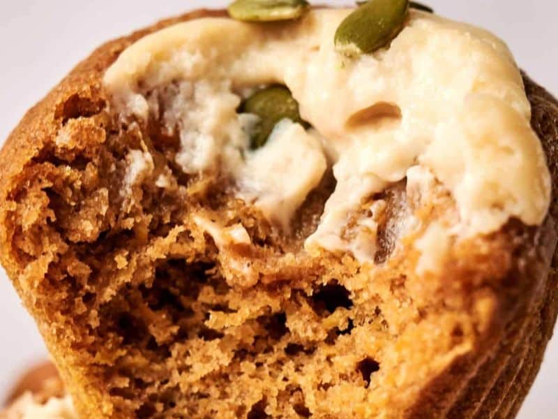 Close-up of a partially eaten muffin with a creamy topping and pumpkin seeds on top.