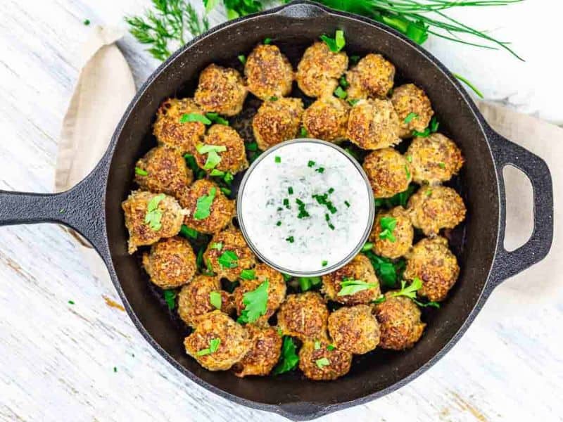A cast iron skillet filled with cooked meatballs garnished with herbs, arranged around a bowl of white dipping sauce topped with chopped chives.