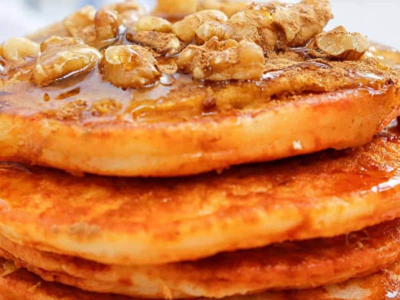 A close-up view of a stack of pancakes topped with walnuts, syrup, and a sprinkle of cinnamon.