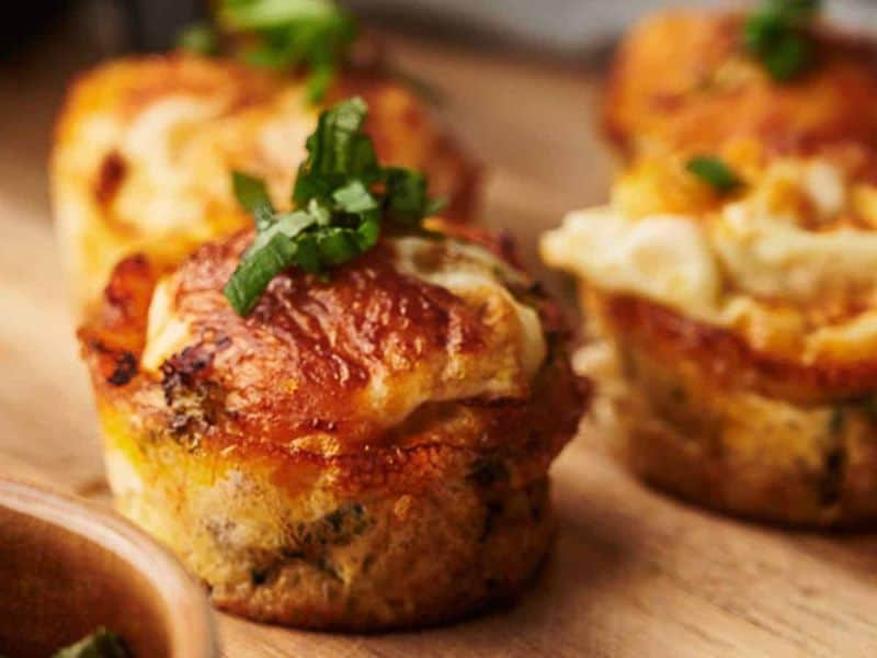 Three savory muffin-like egg bites topped with melted cheese and chopped green herbs, displayed on a wooden surface.