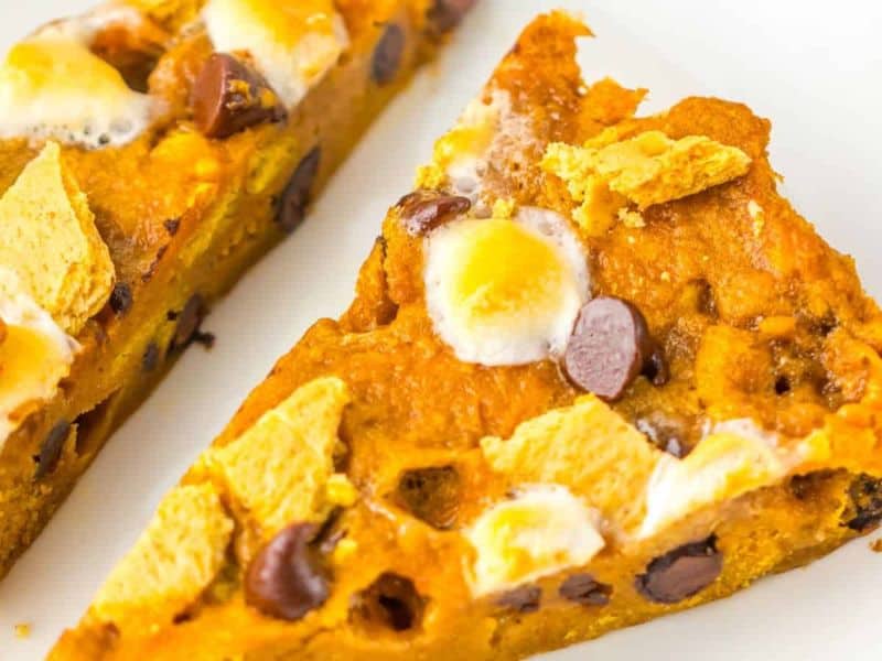 Two triangular slices of dessert bar topped with chocolate chips, mini marshmallows, and pieces of graham cracker on a white surface.