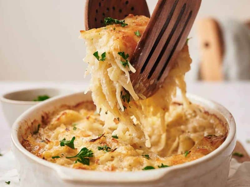 A baked cheesy potato casserole is being served from a white dish with wooden utensils, topped with chopped parsley.
