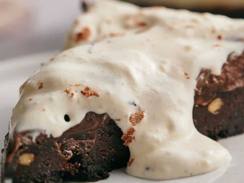 A slice of chocolate brownie topped with a generous layer of creamy vanilla sauce on a white plate.