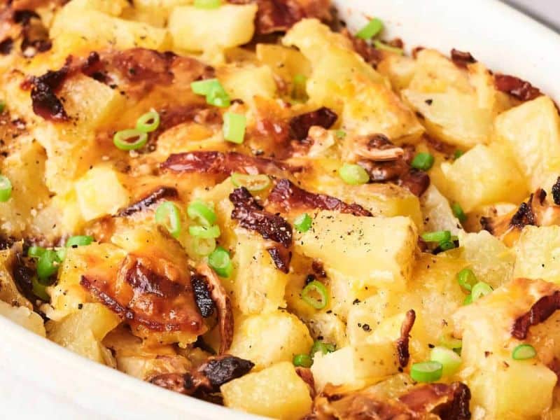 A close-up of a baked casserole dish with chunks of potato, crispy bacon, melted cheese, and garnished with chopped green onions.