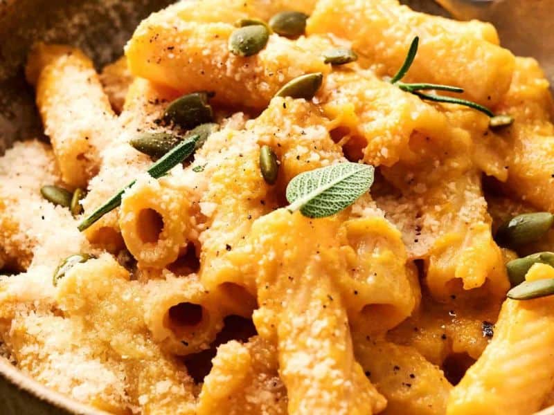 Close-up of rigatoni pasta coated in a creamy orange sauce, topped with grated cheese, pumpkin seeds, and fresh herbs.
