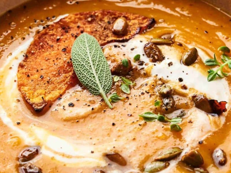 Close-up of creamy pumpkin soup garnished with a slice of roasted pumpkin, fresh sage, thyme, pumpkin seeds, pepper, and a swirl of cream.