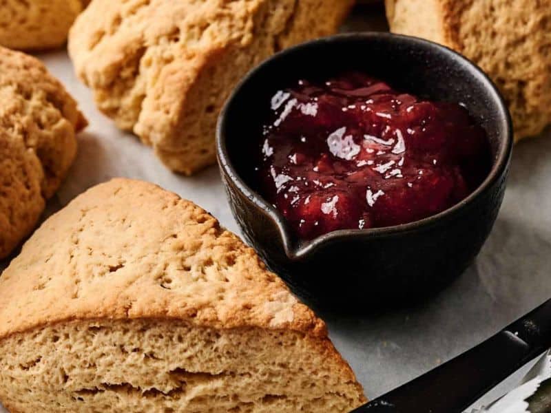 Golden brown scones arranged on parchment paper next to a small black bowl filled with red fruit jam.