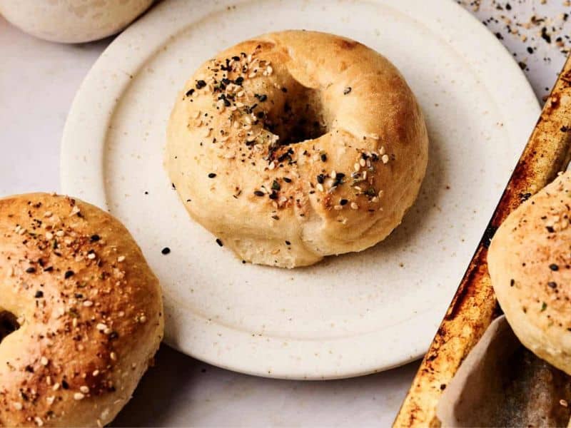 A plain bagel topped with mixed seeds sits on a round white plate next to a baking tray and another bagel.