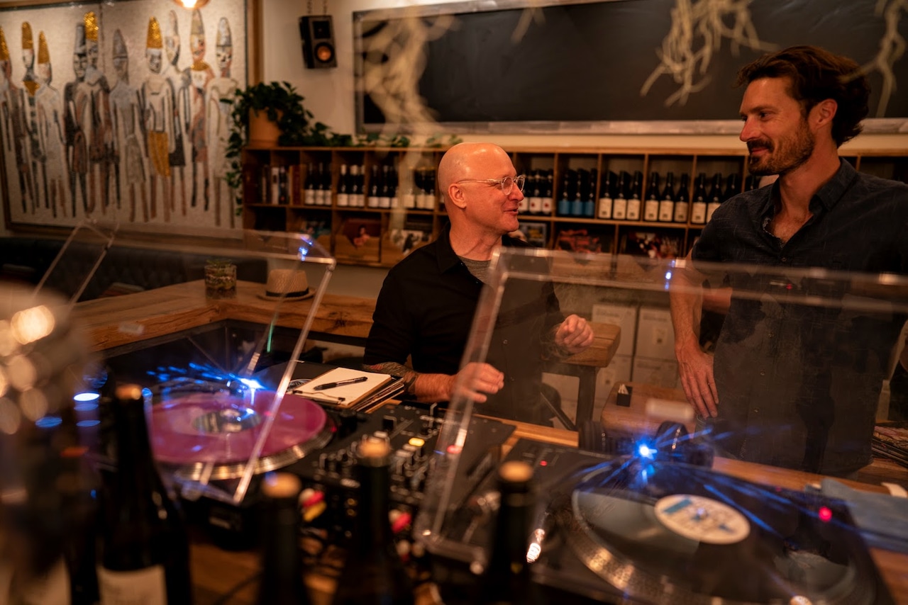 Two men talk in a wine bar in front of two DJ turntables.