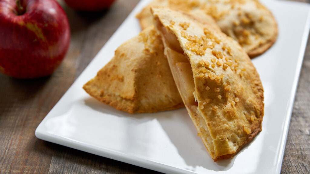 A glamor shot of an apple hand pie sitting on top of another hand pie, on a plate next to an apple.