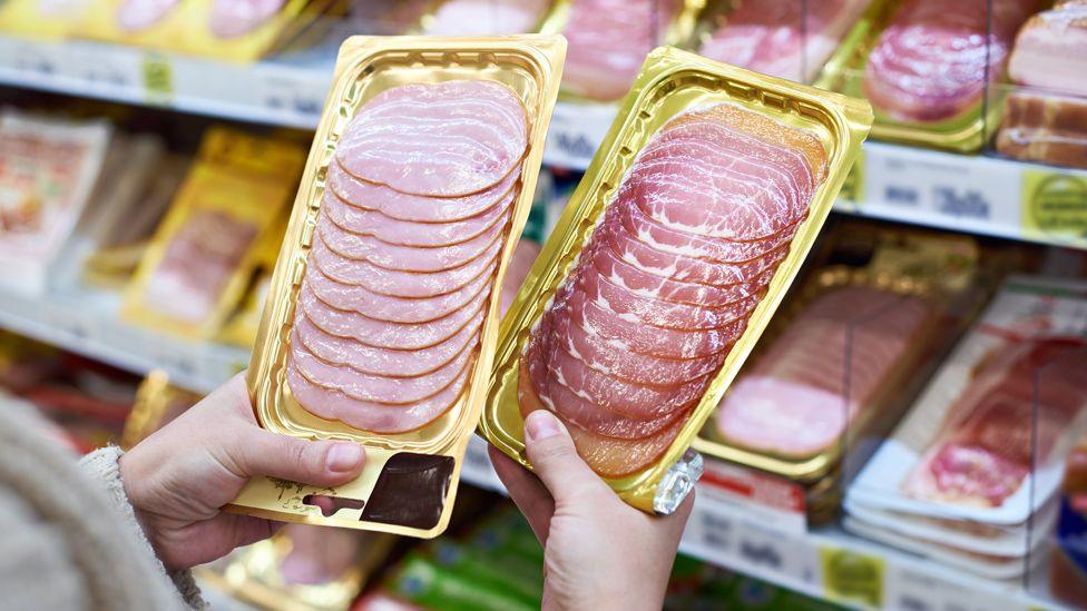 A close up of a womans hands holding two packets of cured meats in a shop