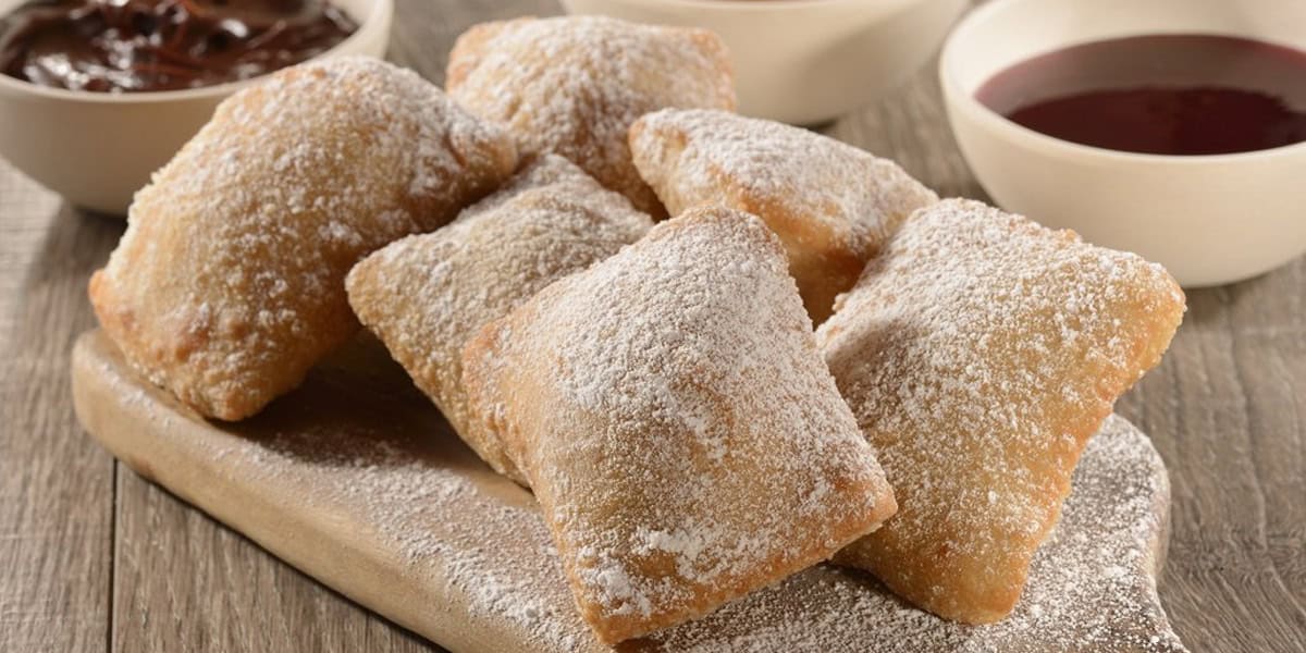 Five golden-brown beignets dusted with powdered sugar are arranged on a wooden board at Les Halles Boulangerie-Patisserie, with bowls of chocolate and berry sauces in the background.