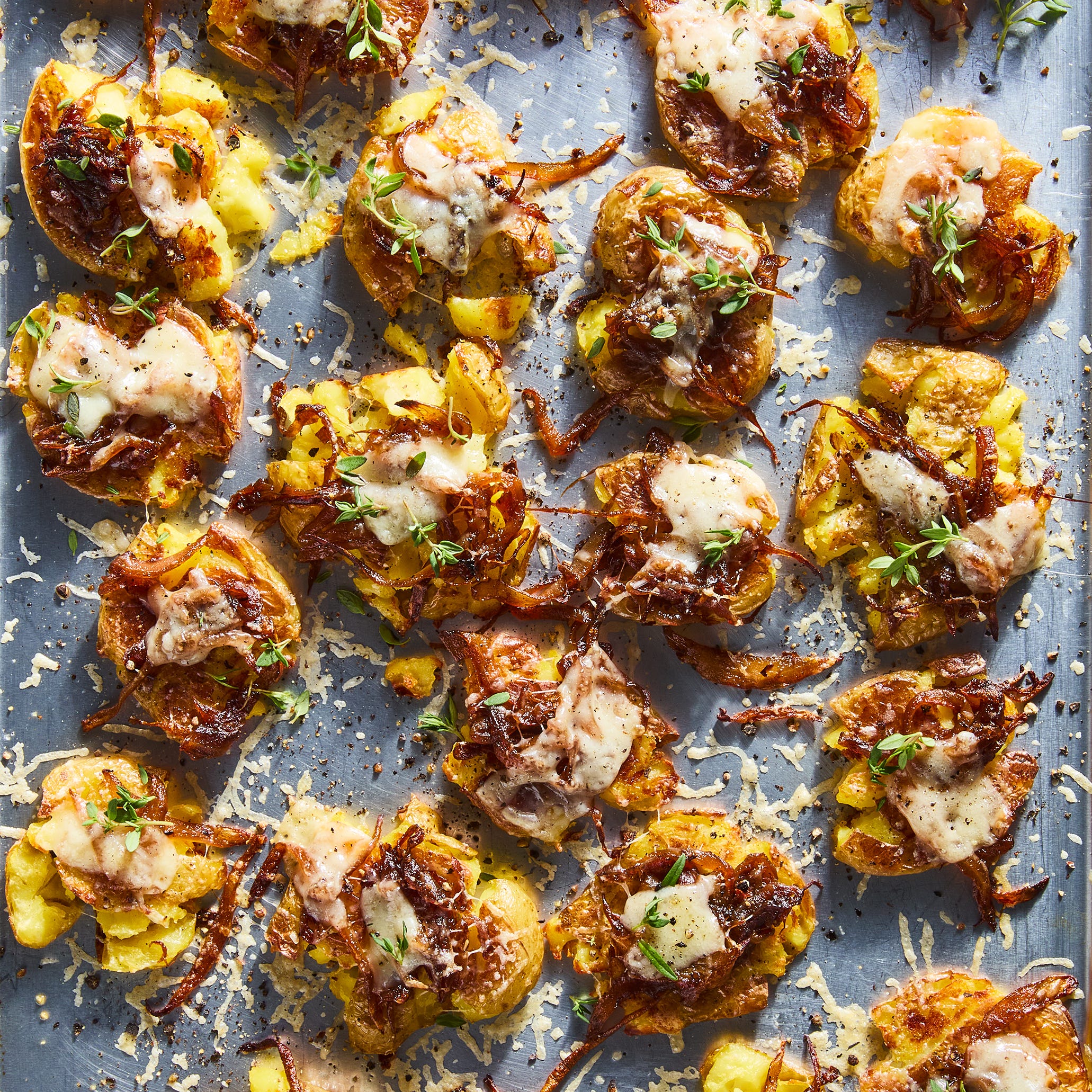 french onion smashed potatoes topped with caramelized onions, gruyere, and thyme on a baking sheet