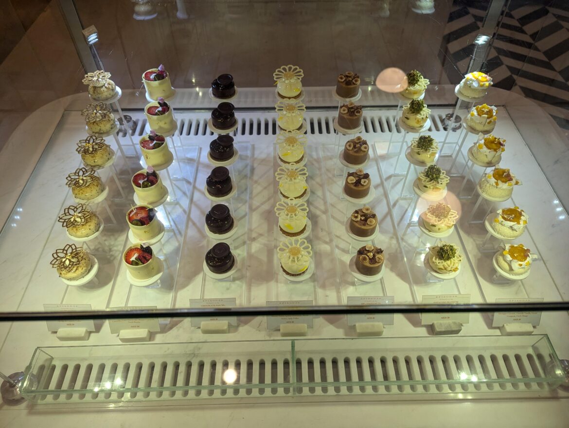 An immaculate display of Petit Gâteaux - including Chocolate Fan 70%, Pistachio Blossom, and 100% Hazelnut cakes at Episodes, Abu Dhabi.