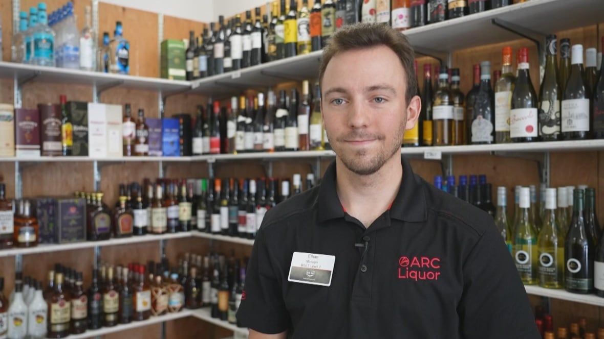 A man wearing a black T-shirt smiles in front of a liquor rack.