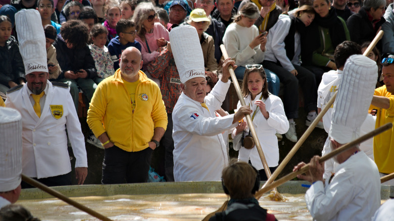 Men in chef's whites using giant wooden spoons stir a giant pot of eggs in front of a crowd