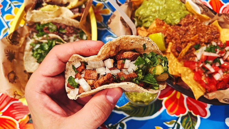 Hand holding taco over plates of Mexican food on table.