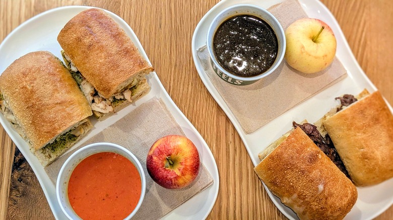 Two sandwiches, each with a cup of soup and an apple, on a wood table