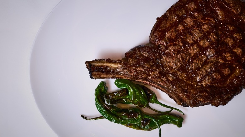 Grilled bone-in ribeye steak on white plate garnished with shishito peppers