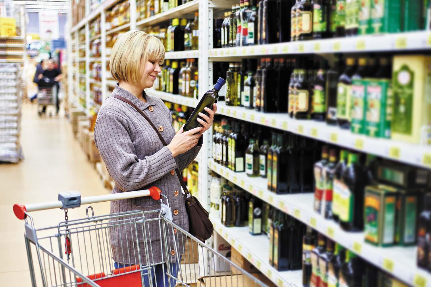 A woman reads the label on a bottle of olive oil while standing in front of shelves holding many different kinds of oil.