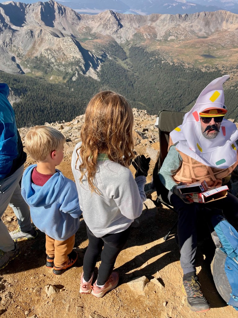 Man in ice cream costume sharing ice cream with children on a mountaintop.
