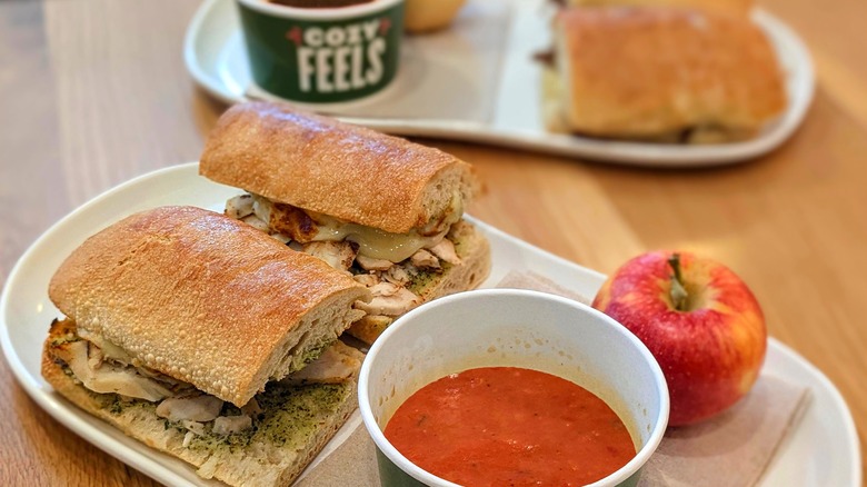 A sandwich on ciabtta bread with soup and an apple and another sandwich in the background on a wood table