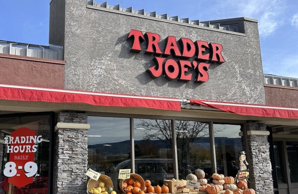The exterior of a Trader Joe’s store with a red sign, stone accents, and a display of pumpkins and gourds outside. A sign in the window shows store hours as 8-9 daily.