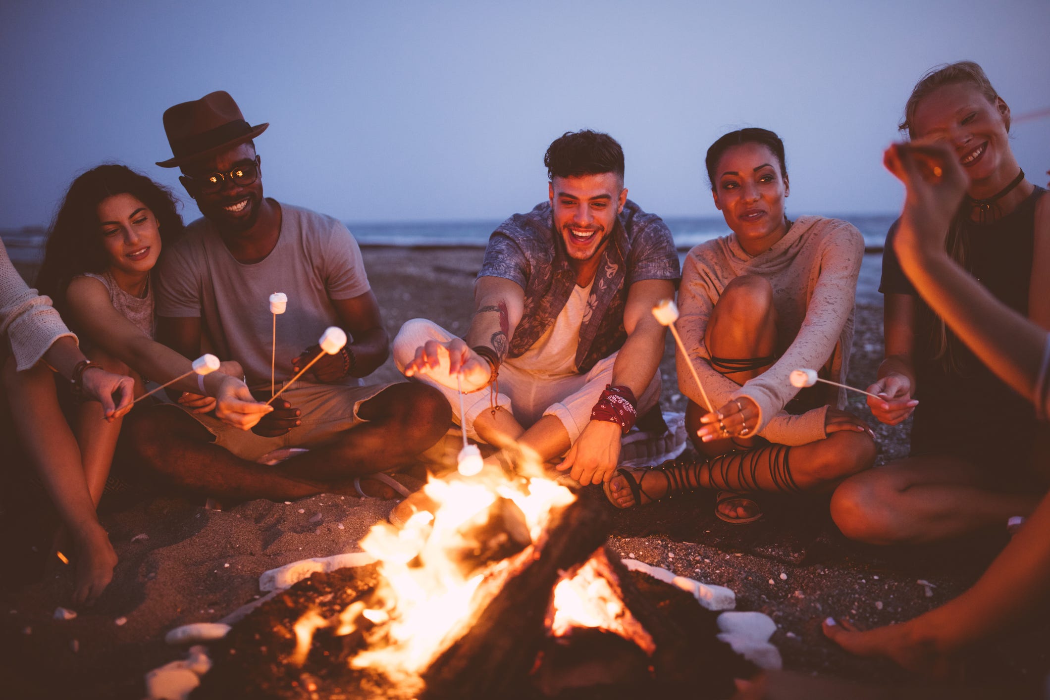 young multi ethnic friends roasting marshmallows on sticks at the beach