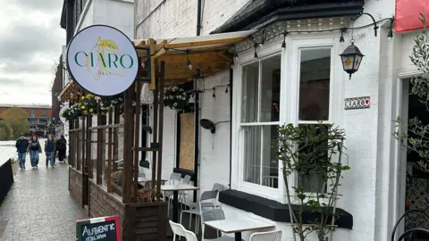 BBC The front of Ciara Italian Deli restaurant. The building is white with windows and black windowsills. One of the windows has a board covering it. There are white tables and chairs outside and people can be seen walking on the canal path next to the river.