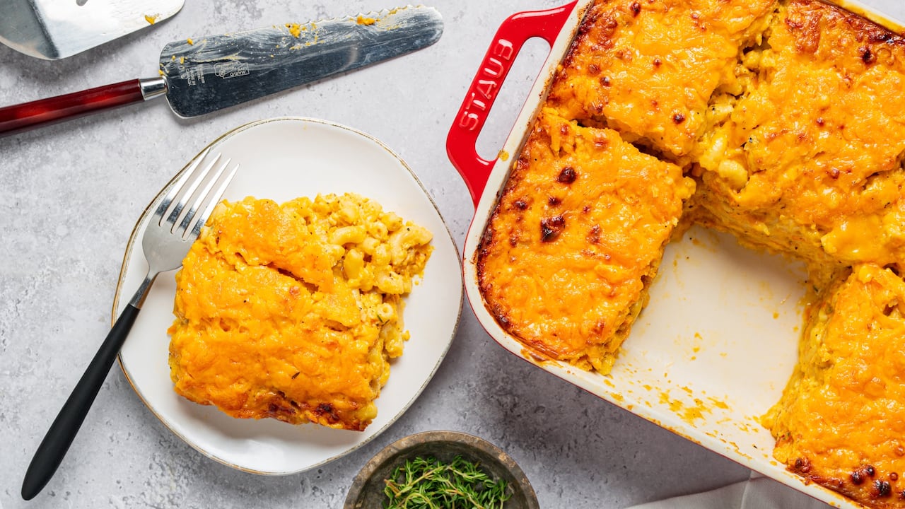 Orange, cheesy macaroni pie in a rectangular baking dish on a counter. A slice of it is sitting on a plate next to the dish. 