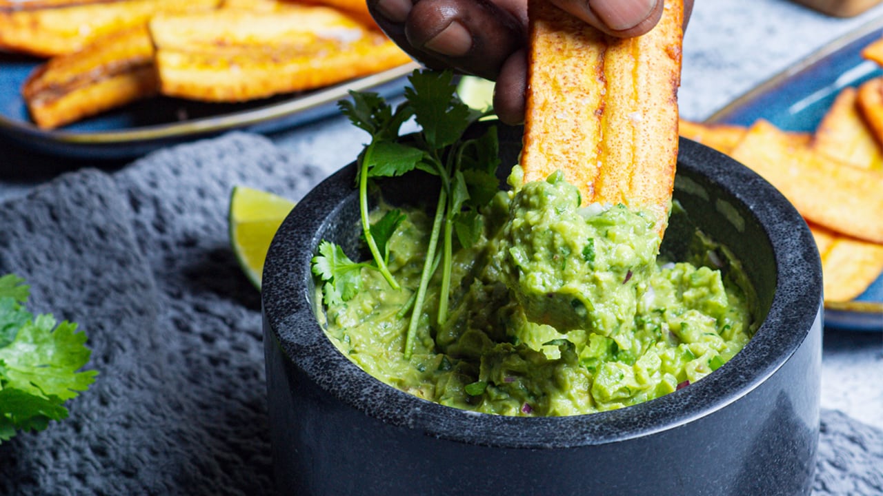 Closeup on a hand using a fried plantain to scoop guacamole from a grey bowl.