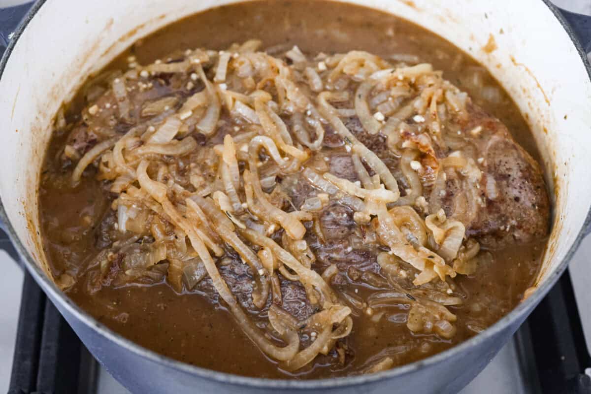 This French Onion Pot Roast is pure comfort food! Slow-cooked to perfection, the roast becomes melt-in-your-mouth tender as it simmers in a rich, savory gravy infused with the deep, caramelized flavor of onions. Close up shot of the french onion pot roast with caramelized onions and broth around it ready to go in the oven.