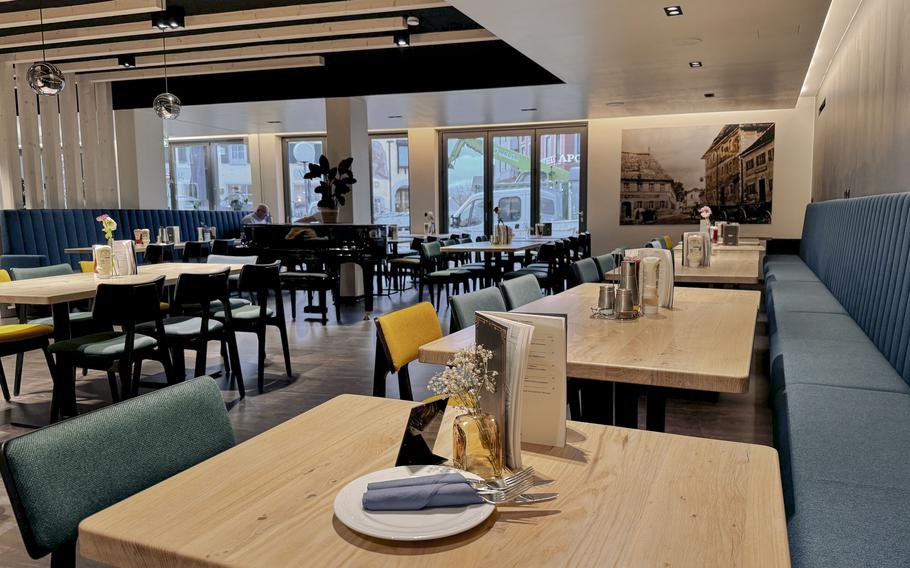 Spacious main dining room with floor-to-ceiling windows overlooking the street, modern seating in teal and black, a grand piano and contemporary artwork on walls.