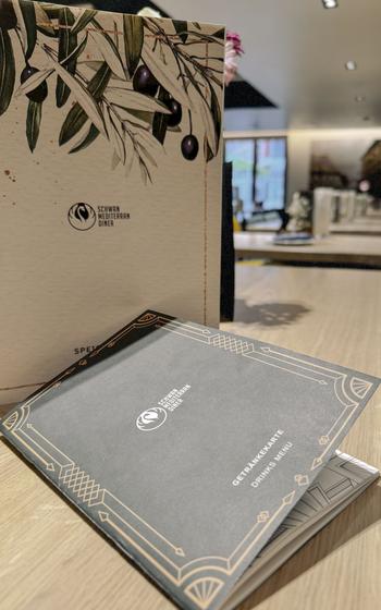 Schwan Mediterranean Diner menu and drinks menu displayed on a wooden table with an olive branch illustration on the cover.